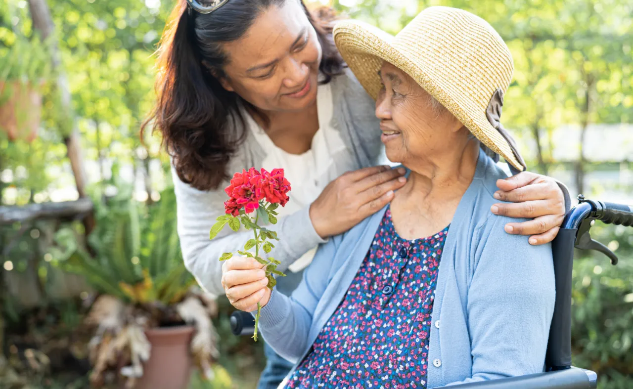 Caregiver presenting red flower to elderly woman in wheelchair outdoors