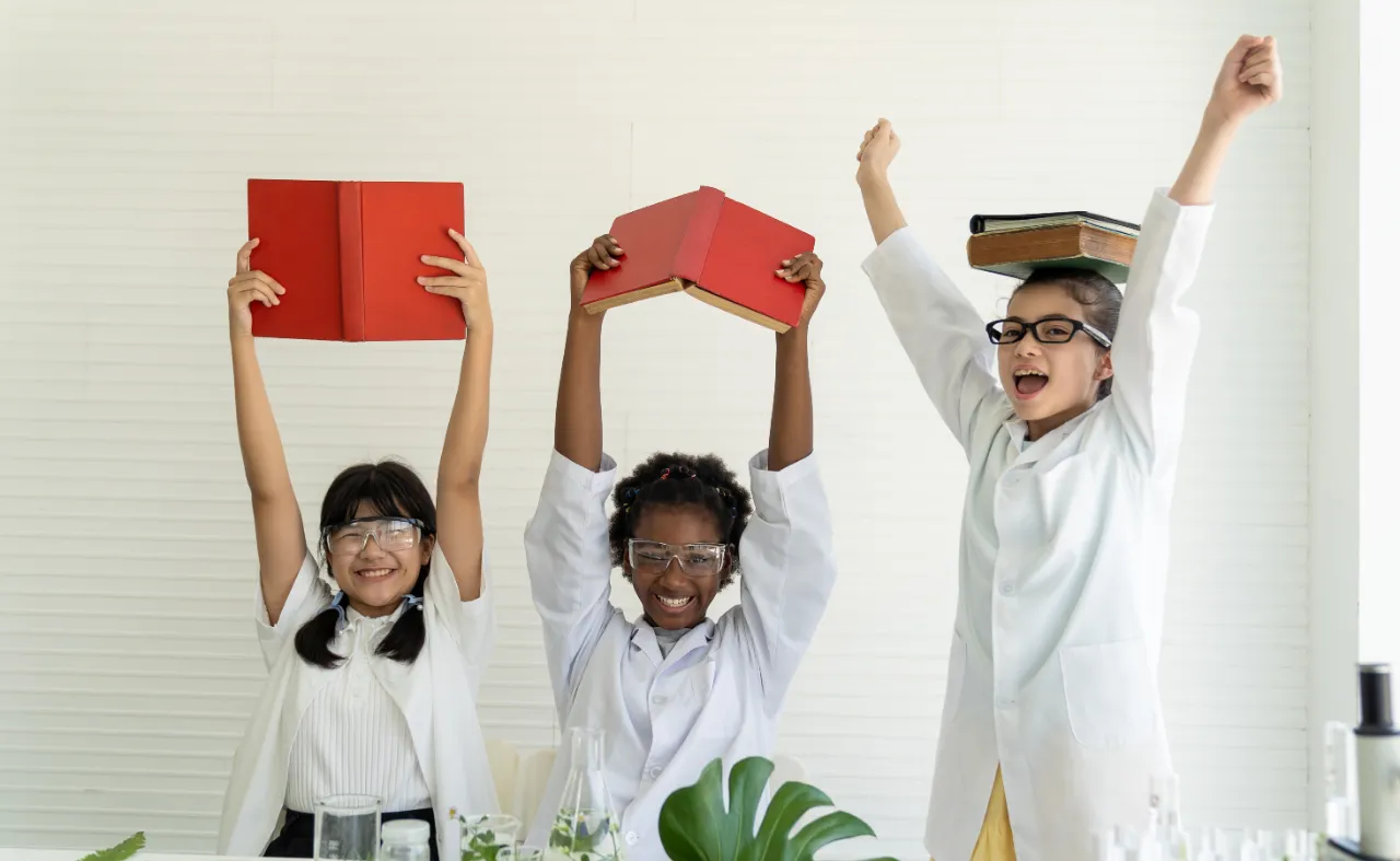 Three excited girls in lab coats celebrating with books